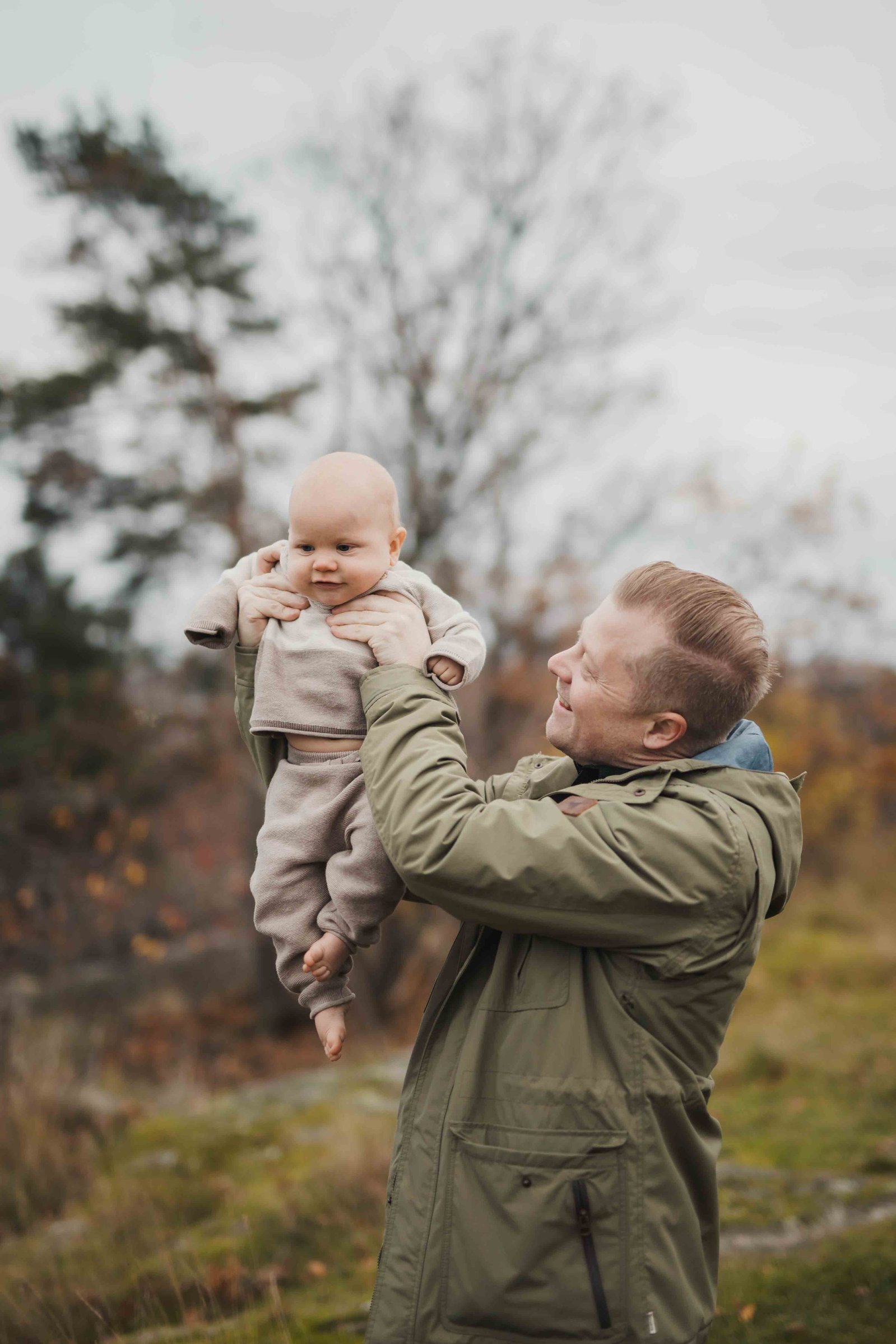Pappa i grön jacka lyfter upp sin son högt i luften, bebisen ler förnöjt - kläd i beiga varma kläder.