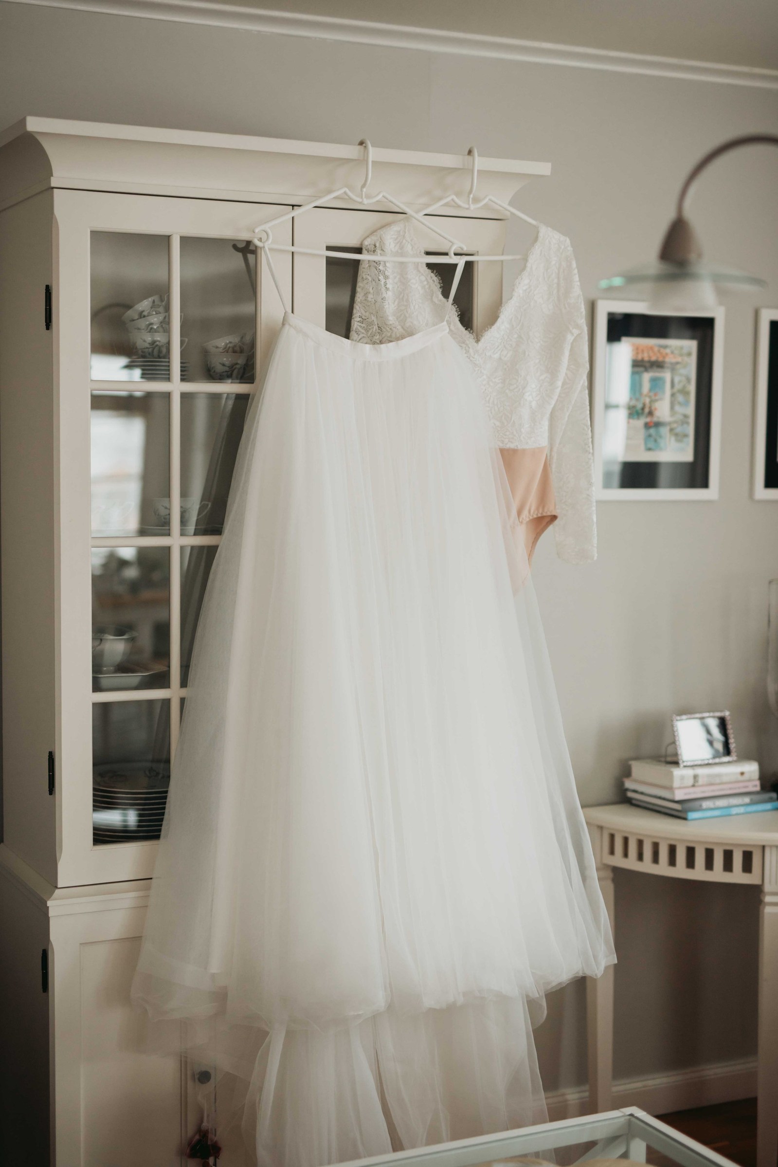 Brudens vita tyllkjol hänger på vit galge tillsammans med den vita spetstoppen i vitrinskåpets framsida av glasparti - i ett vardagsrum med böcker och tavla på väggen.