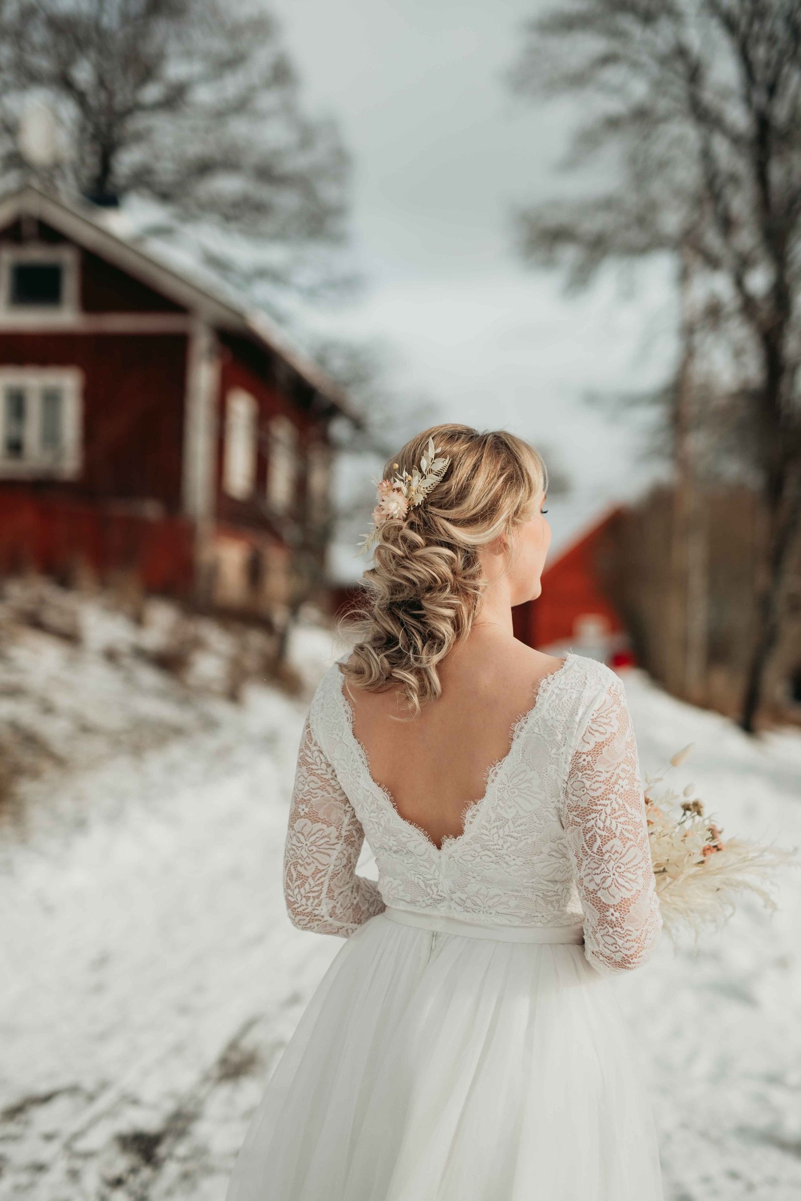 Brud med vit spets top och vit tyllkjol står utomhus i snön på landet i bakgrunden faluröd stuga och träd. Håret är uppsatt i lockar med hårsmycken i rosa torkade blommor.