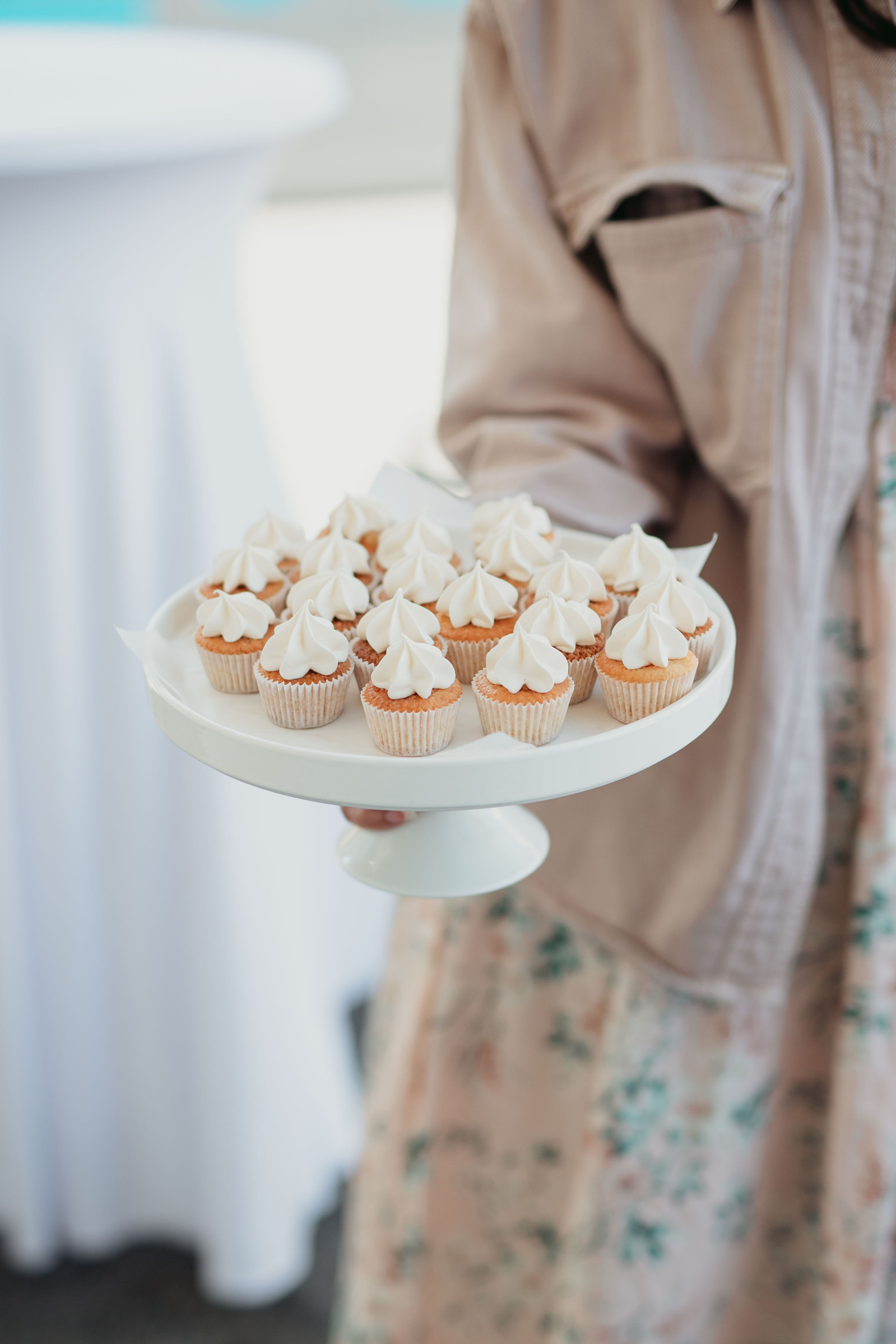 Runt vitt kakfat med fot med cupcakes med vitt topping - hålls med ena handen av kvinna med rosa jacka och blommig kjol.