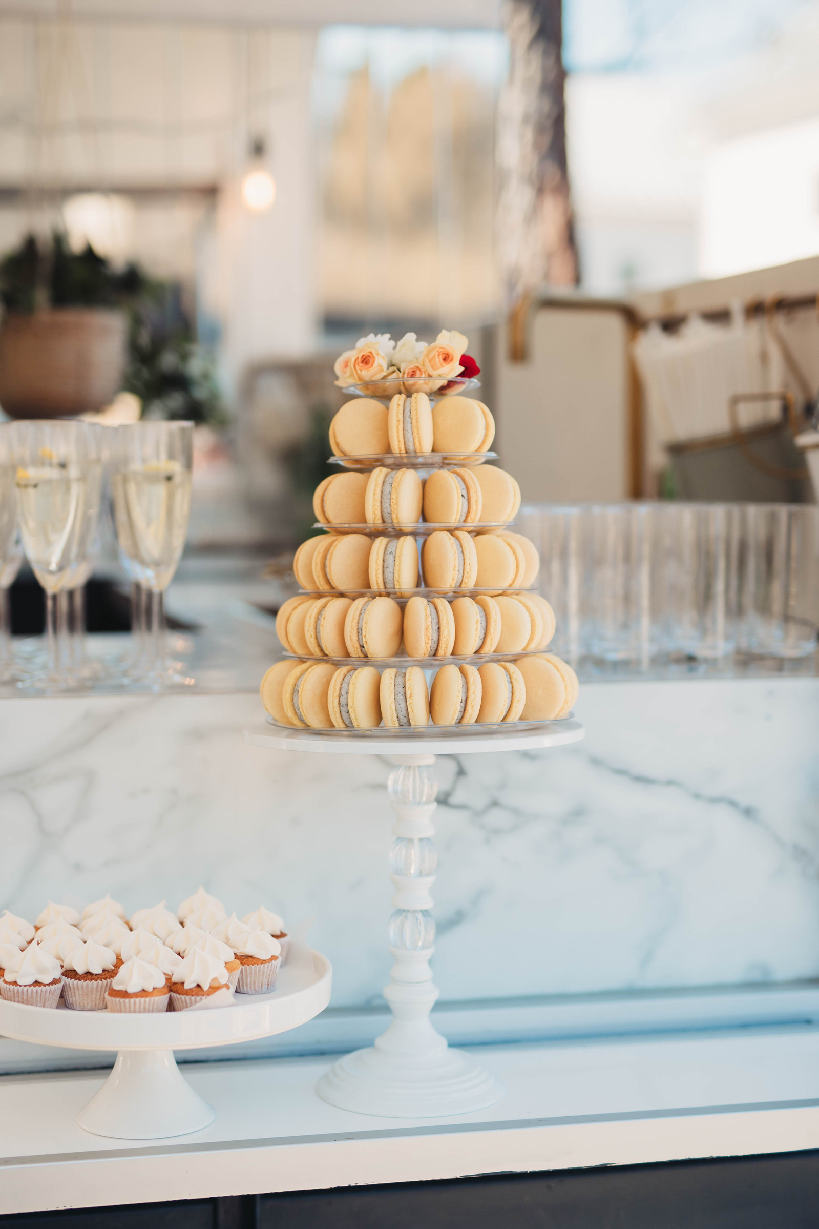 Gula macarons i pyramid på vitt kakfat med blommor på toppen.  I bakgrunden bar och glas för gäster, till vänster ett lågt vitt kakfat med cupcakes med vit topping.