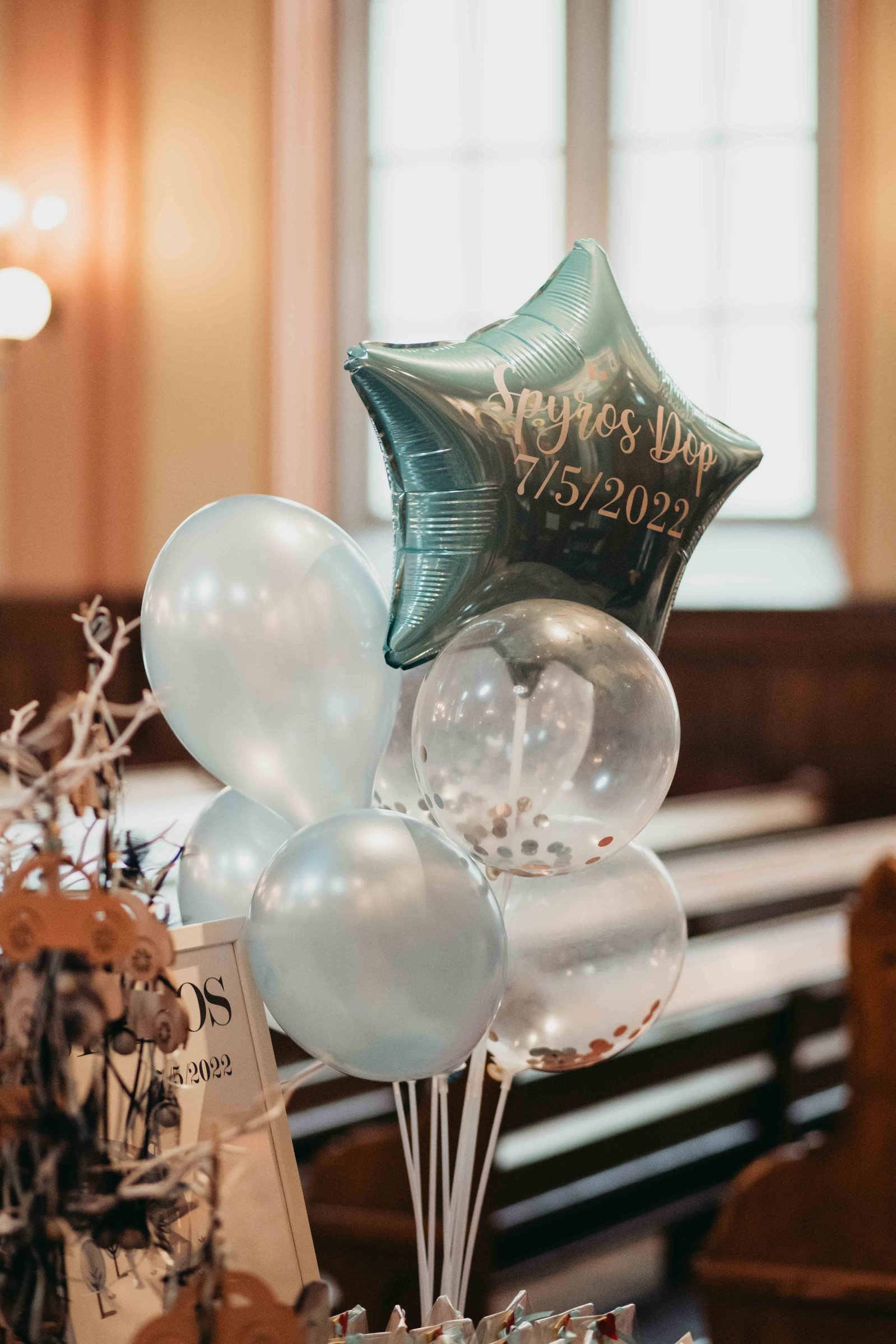Runda ballonger i ljusblått och genomskinligt material samt en i form av en stjärna med tryck med namnet Spyros  7/5/2022 uppställt  framför bänkraderna i grekiska kyrkan.