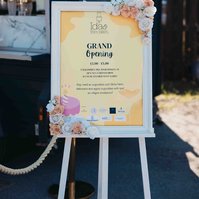 Vit stafli med gul skylt och vit ram dekorerad med blommor och texten " Grand Opening" står utomhus.