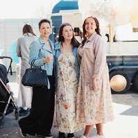 3 kvinnor poserar på en invigning utomhus, en klädd i jeansjacka, svart byxa och väska i handen, hon i mitten med blommig lång klänning och kvinnan längst ut rosa jacka och blommig klänning.