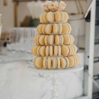 Gula macarons i 5 våningar och toppen med  blommor står på en ställning och barvagn med cup cakes.