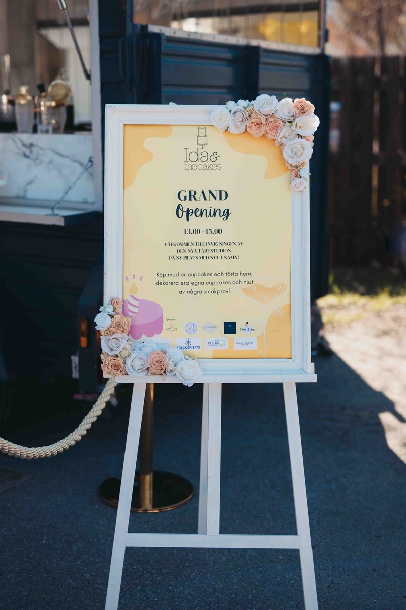 Vit stafli med gul skylt och vit ram dekorerad med blommor och texten " Grand Opening" står utomhus.