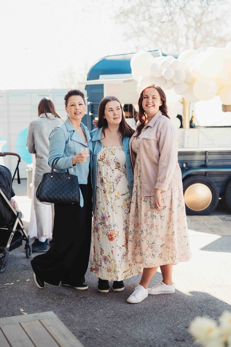 3 kvinnor poserar på en invigning utomhus, en klädd i jeansjacka, svart byxa och väska i handen, hon i mitten med blommig lång klänning och kvinnan längst ut rosa jacka och blommig klänning.