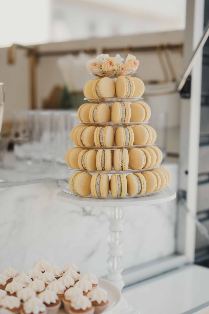 Gula macarons i 5 våningar och toppen med  blommor står på en ställning och barvagn med cup cakes.