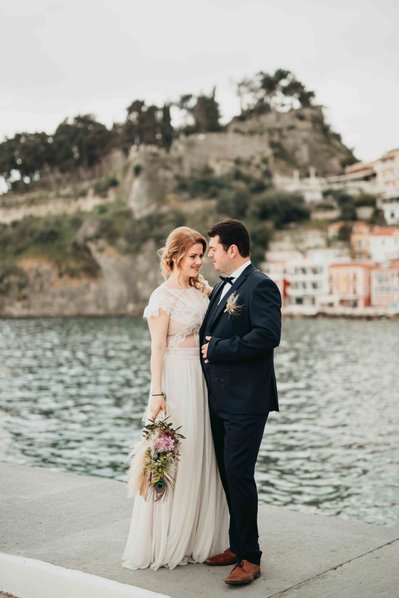 Brud i lång vit klänning  och budbukett står tätt ihop med brudgum i mörkblå kostym vid hamn i Grekland. I bakgrunden grekiska husfasader samt venetianska borgen på toppen av en kulle.