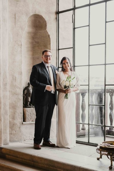 Brudpar står framför ett panorama fönster i glas inne i Stockholms stadshus. Bakom dem stenmur i beige färg. Han i blå kostym och hon i vit klänning och brudbukett i handen.