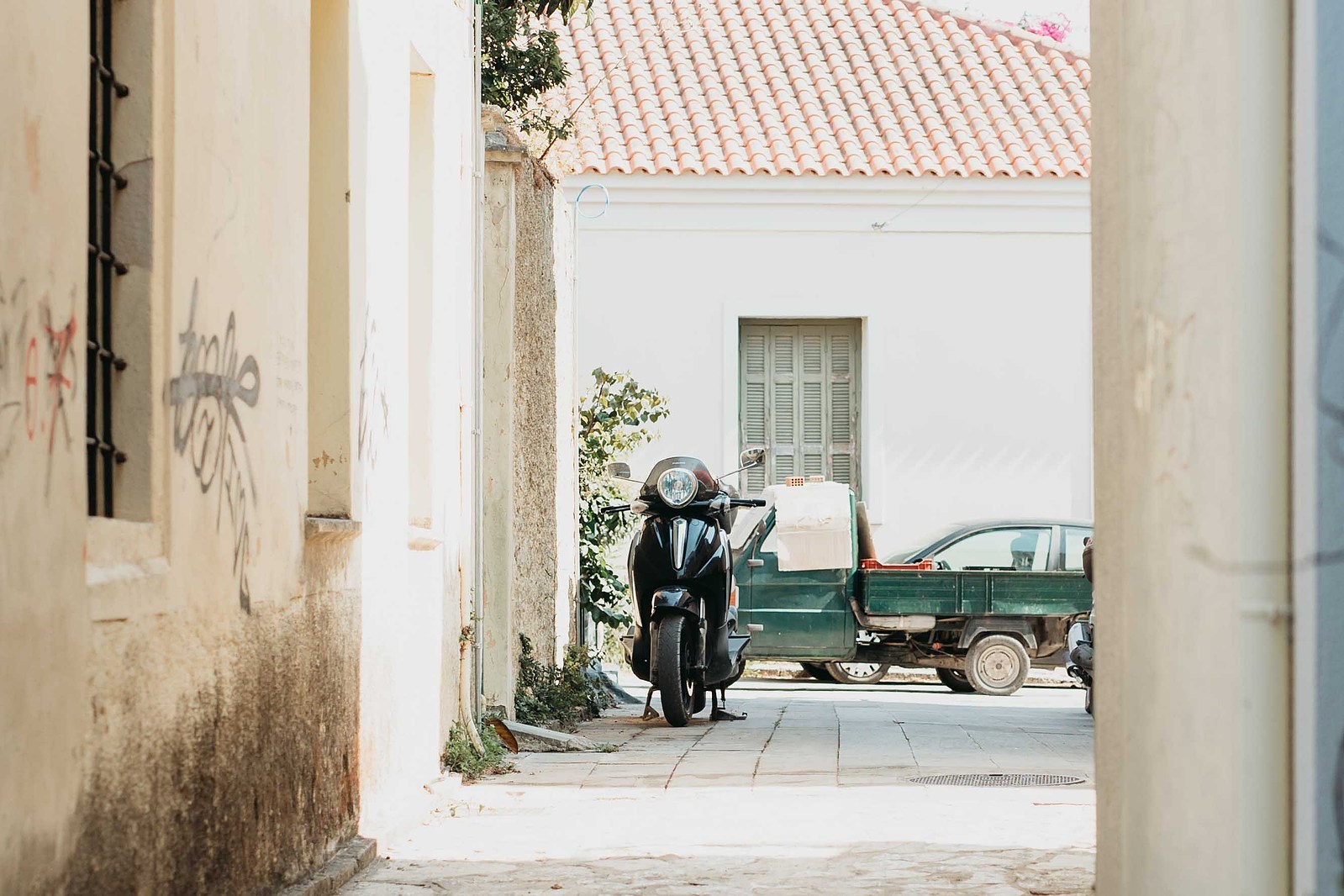Vespa parkerad på en gränd i Greklan. Gul fasad och fönster till höger, bakom vespan bil med flak och fönster med luckor i ljusgrön färg.