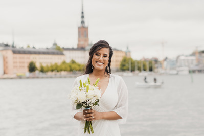 Glad brud i vit klänning och brudbukett i handen med vita blommor. I bakgrunden Riddarholmens byggnader och vattnet i Mälaren i Stockholm.