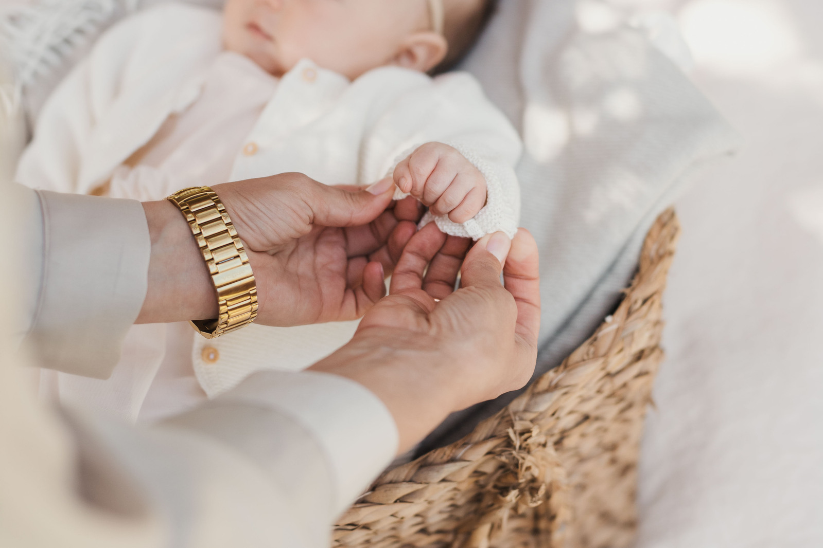 En kvinna håller sin dotters babyhand mellan sina händer. Hon bär en klocka med  guldrem. Baby klädd i rosa och vit kofta,  ligger i en flätad korg.