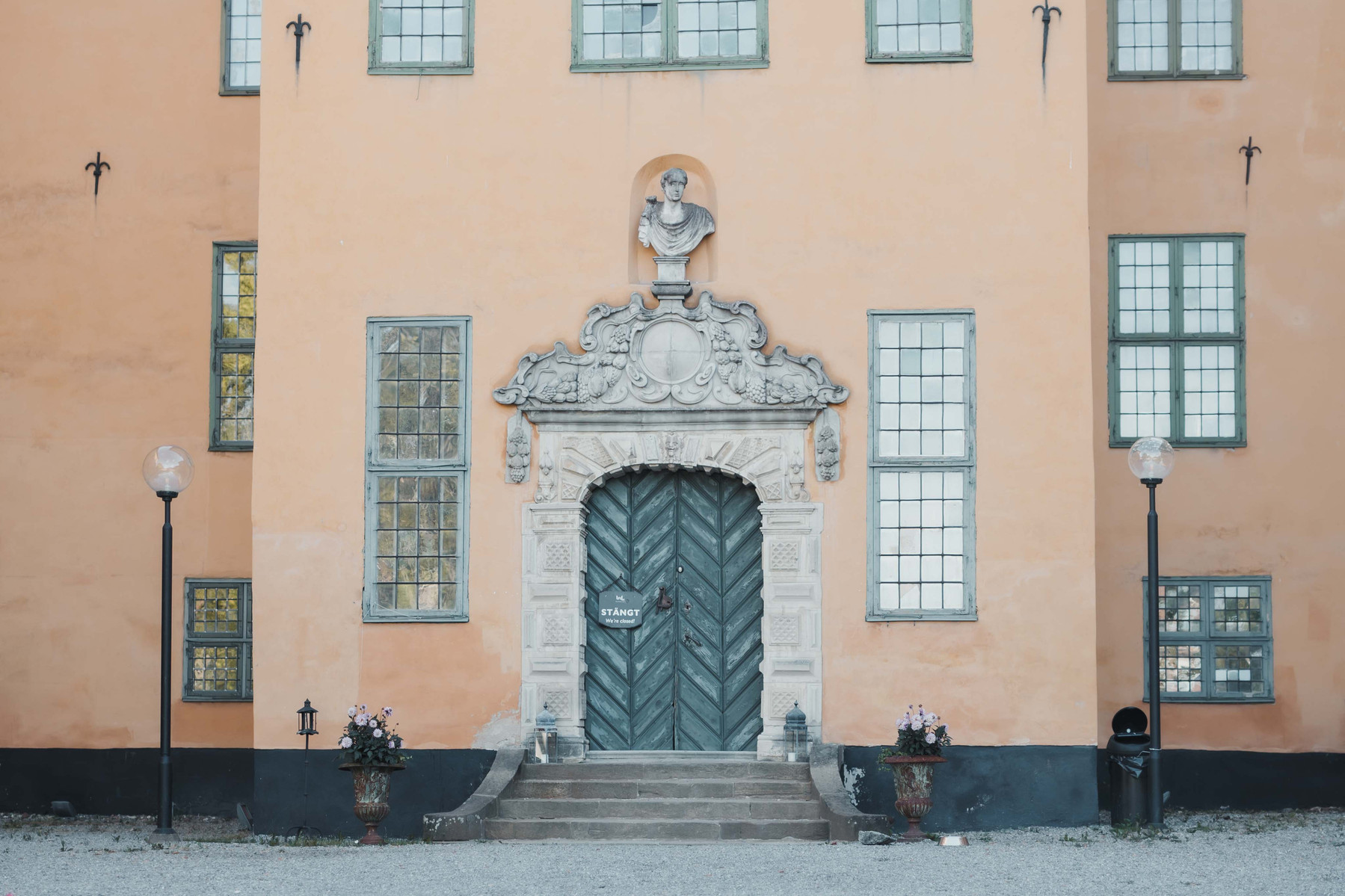 Entrén till slott med gul puts byggt på 1500 talet. Grön dörr med ornament och byst ovanför dörren. Fönster på var sida om dörren, 3 ovanför och flera andra på övrig fasad. Trappa med blomkrukor och lampstolpar i svart runt entrén.