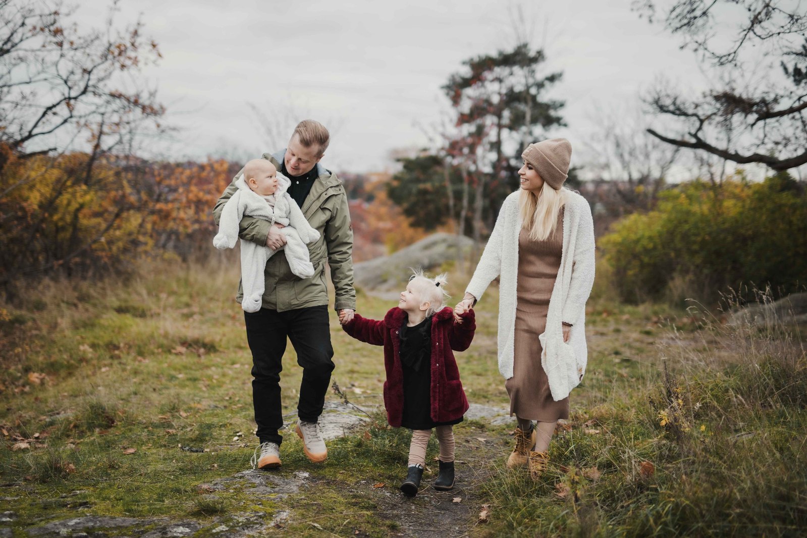 Familj gåendes i naturen med en pappa medvbebis i famnen, en dotter i röd kappa och mamma i vit kofta och mössa. Dottern håller i föräldrarnas händer.