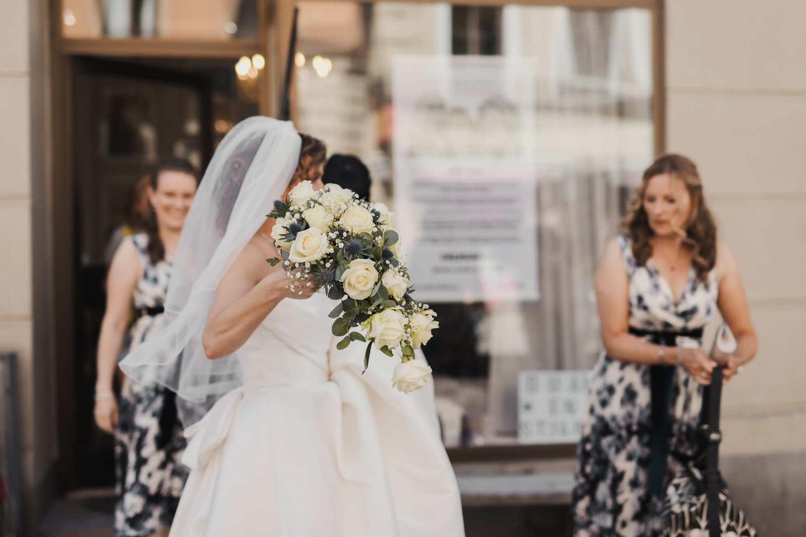 Kvinna i vit brudklänning och vit slöja håller upp sin brudbukett med gula rosor och tistlar utanför en salong med skyltfönster i stan medan hennes tärnor i blåvita klänningar hjälper henne med klänningen.