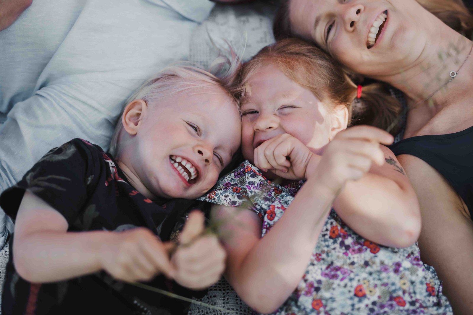 Barnfamilj ligger på en blårandig filt och skrattar tillsammans. Pojke med blont hår blå tröja med palmer, flicka med småblommig sommarklänning. Mamma med ärmlöss blå topp och pappa med ljusblå linneskjorta.