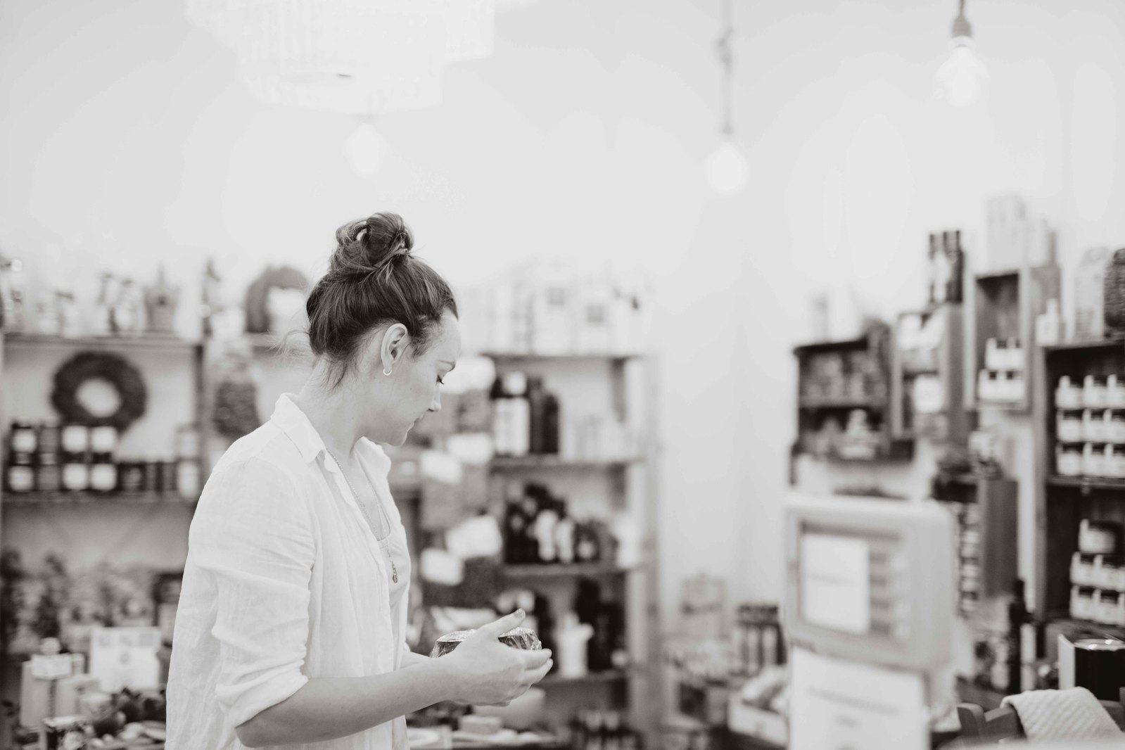 Kvinna i vit skjorta och mörkt hår i uppsatt i en knut håller en skål handen, i bakgrunden butikshyllor med varor.