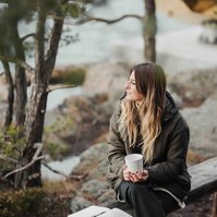 Kvinna med långt cendré färgat hår och grön jacka sitter på en bänk i skärgården och håller i en vit mugg.i Bakgrunden tallar och stenhällar.