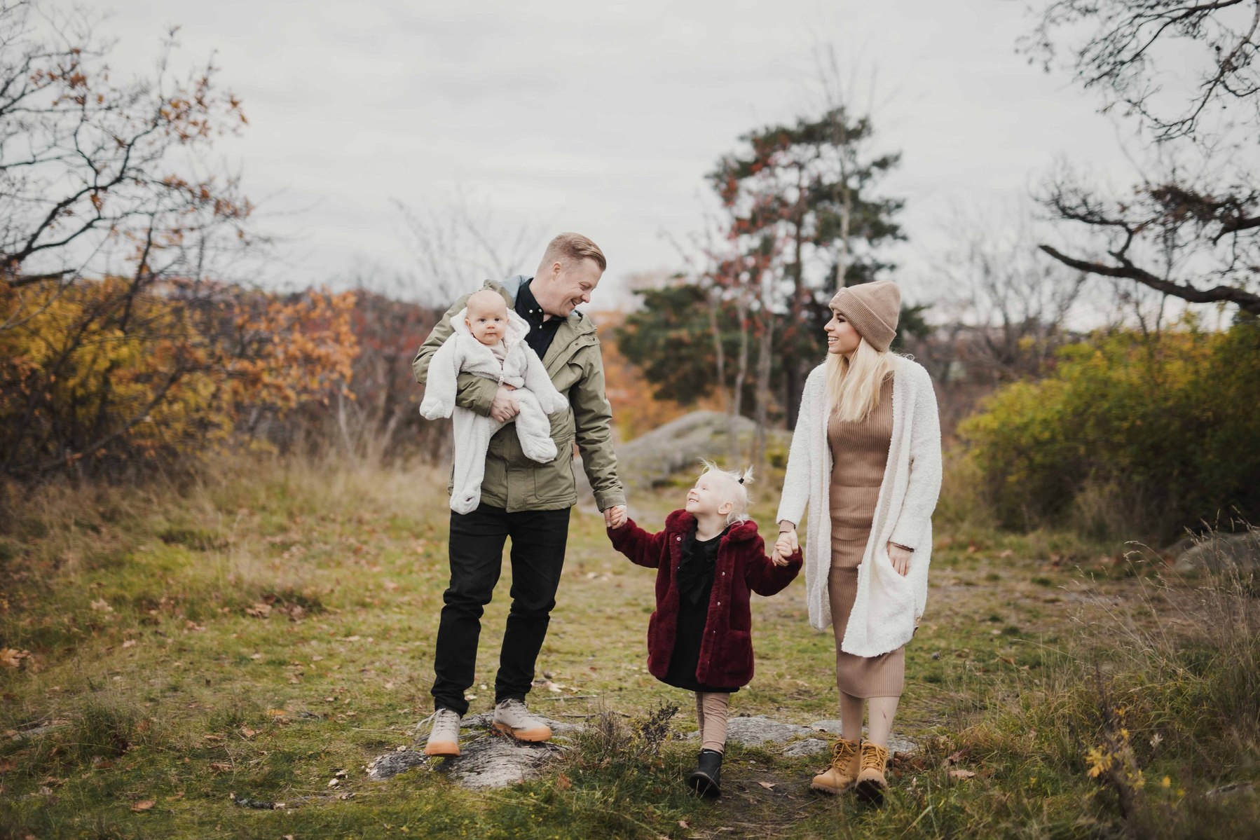 Familj med pappa som håller bebis famnen håller det stora barnet i handen på promenad på hösten i Stockholm. Mamma i vit kofta och beige stickad mössa håller sin dotter handen på andra sidan.