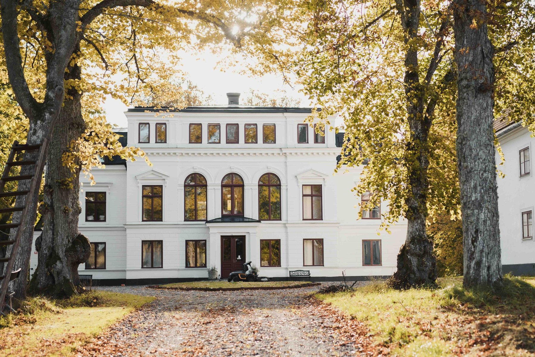 Vit herrgårdsbyggnad i flera våningar omgiven av lövträd. Grusväg och liten rondell med traktor och parkbänk  framför entren.