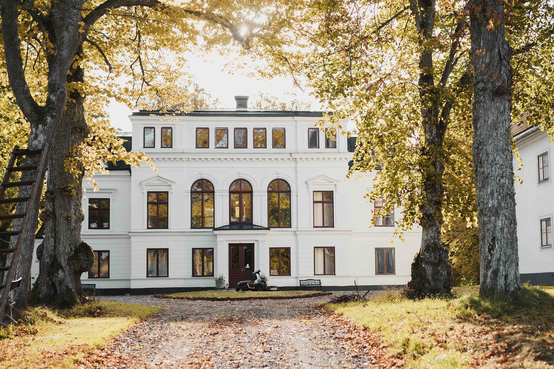 Vit herrgårdsbyggnad i flera våningar omgiven av lövträd. Grusväg och liten rondell med traktor och parkbänk  framför entren.