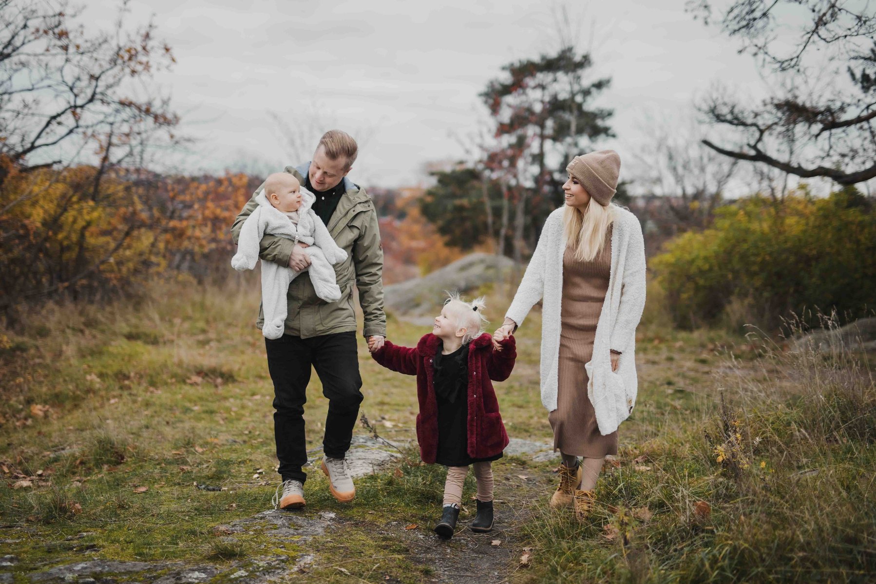 Familj med 2 barn på promenad. Pappa håller en bebis i vitt overall i famnen och håller in dotter som går med den andra. Dotter med röd rock och blont hår håller ssin mamma med den andra handen.