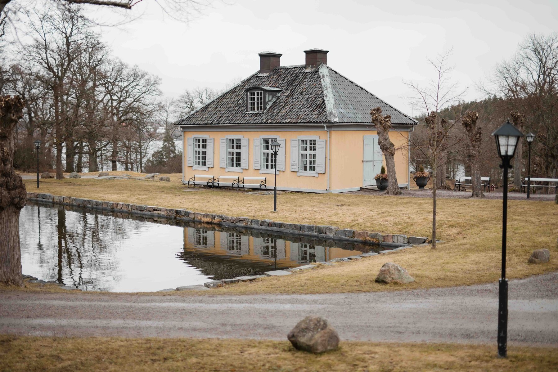 Trä paviljong i gul träpanel och svart tak, omgiven av vattendamm träd och gräsmatta.