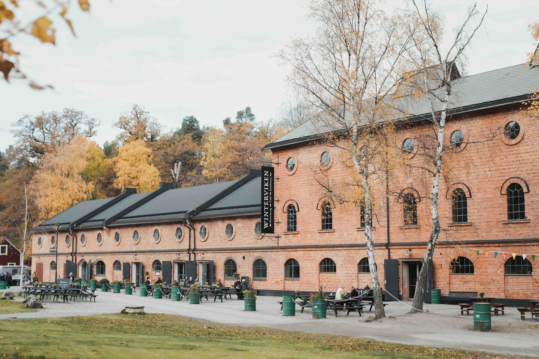 2 tegelbyggnader intill varandra med flera våningar, svart tak, entre med uteplats med 2 björkar och bord för fika. Bakom lövträd i höstfärger, framför fika platsen gräsmatta.
