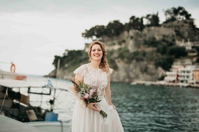 Brud i vit klänning med brudbukett i handen håller i sin klänning vid en hamn, bakom båt med grekiska flaggan. I bakgrunden vattnet i hamnen och berg med en venetiansk borg bakom träden.