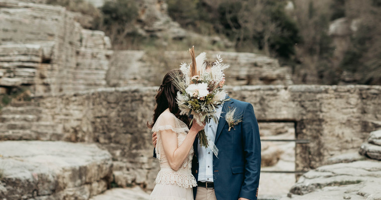Par i elopement fotografering står utomhus bland natur pooler i nordvästra Grekland. Hon håller brudbuketten framför dem, han klädd i blå kavaj, hon i beige klänning med spets. Han håller i med sin hand på hennes rygg.