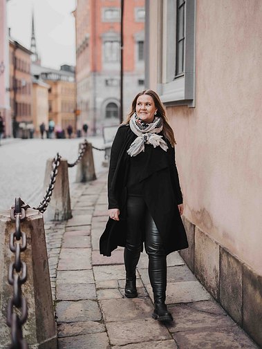 Kvinna med långt hår klädd i svarta byxor och kappa, vit sjal går utomhus på stan. I bakgrunden rosa fasader från stadsmiljö.