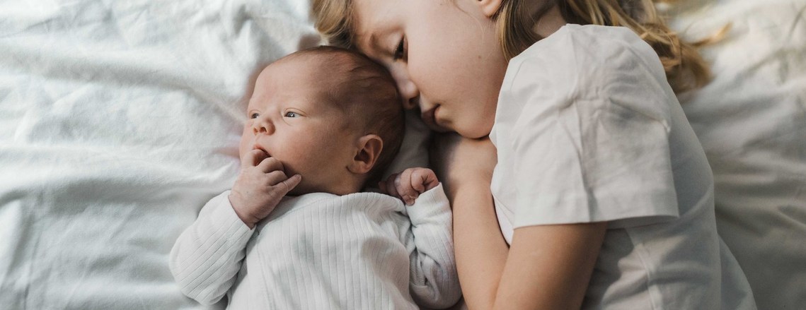 Nyfödd bebis  i vitt body ligger tätt med sin storasyster i t-shirt sängen med vitt skrynkligt lakan.