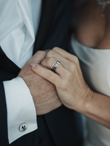En man och en kvinna håller varandras hand tätt tillsammnas - hennes hand ovanpå så att vigselringarna syns,  en med slät ring med vit guld och den andra med stenar. Han har manschettknapp till vita skjortan, hon i vit klänning.