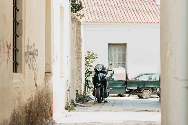 Vespa parkerad på en gränd i Greklan. Gul fasad och fönster till höger, bakom vespan bil med flak och fönster med luckor i ljusgrön färg.