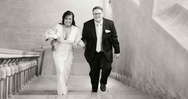 Par går uppför trappan i Stadshuset i Stockholm på väg till Ovalen för vigsel. Hon i vit klänning och brudbukett i handen, han i glasögon, blå kostym och slips.
