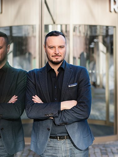 Man i blå kavaj, skjorta och jeansbyxor. lutar. sig med axeln mott. en glasfasad utanför en entre.