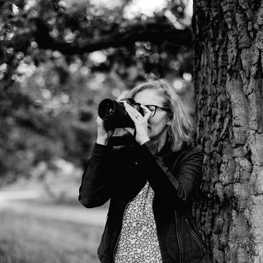 Kvinna i page och glasögon fotograferar med kamera utomhus under ett träd. Klädd i mörk jacka och småblommig blus i vitt och svart.
