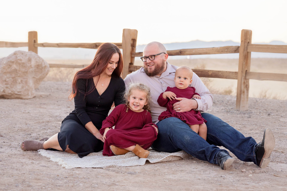 Family posing for a photography session. The family is sitting on a blanket in front of a desert landscape and they are laughing and smiling
