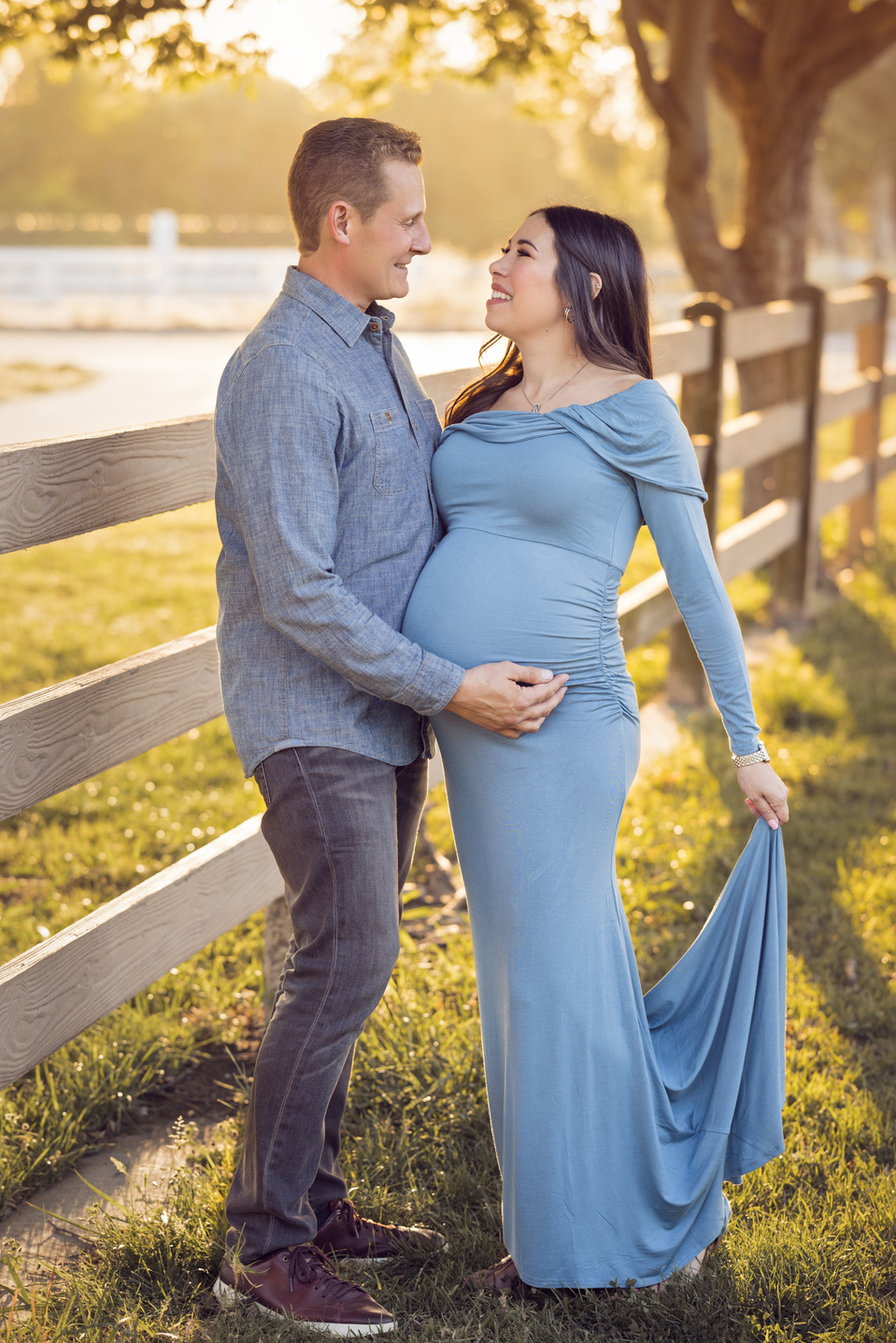 Husband and wife posing for outdoor maternity photoshoot session. Photo is taken at golden hour and the sun is glowing from behind. The couple is standing on the grass in front of a large tree and a wooden fence