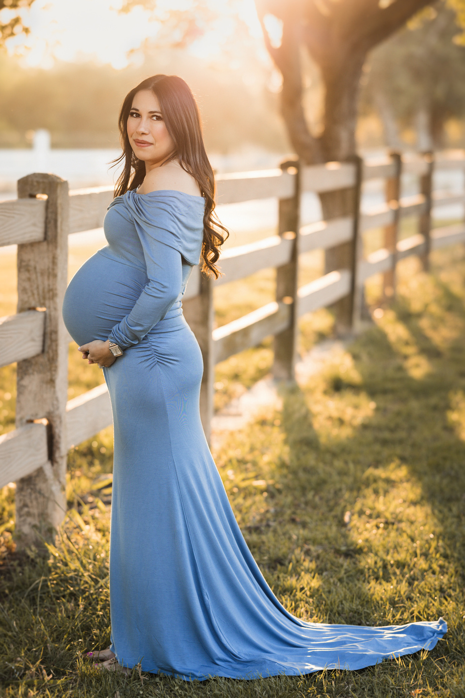 Outdoor maternity session at golden hour. The sun is glowing from behind the pregnant woman. She is standing on a grassy area in front of a big tree and a wooden fence