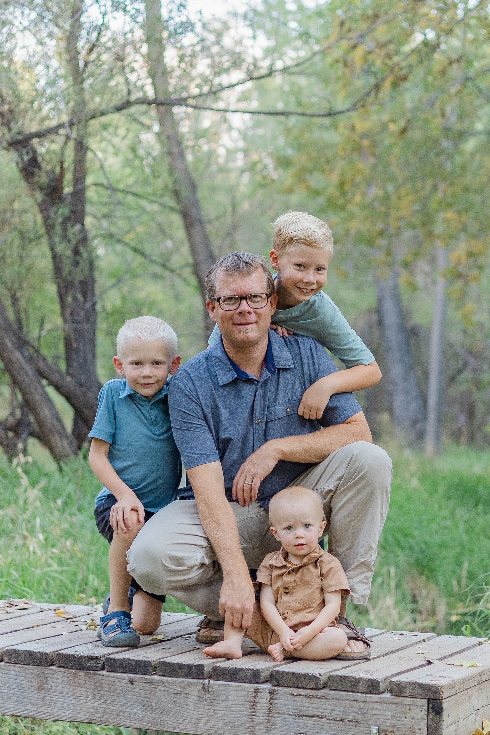 A father is hugged by his two boys while the baby is safe at his feet. The Lens Lady loves to capture connection in her family photography.