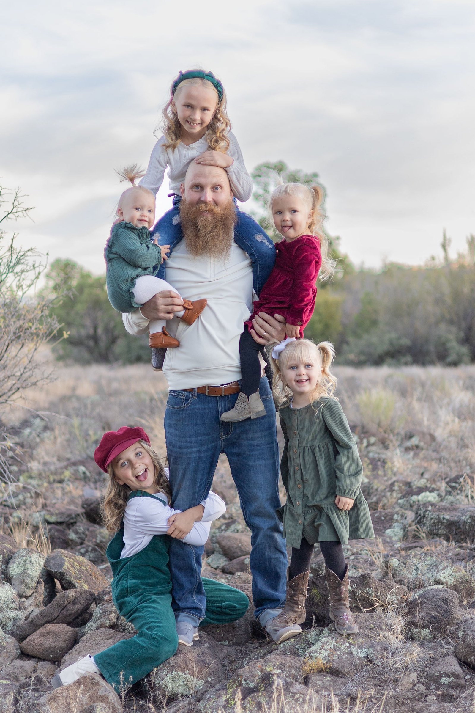 A dad stands smiling in the midst of his 5 girls. One girl is on his shoulders, he has a daughter in each arm, another clings to his leg, and the last daughter stands by his side. Family portraits can be fun and playful with the Lens Lady.