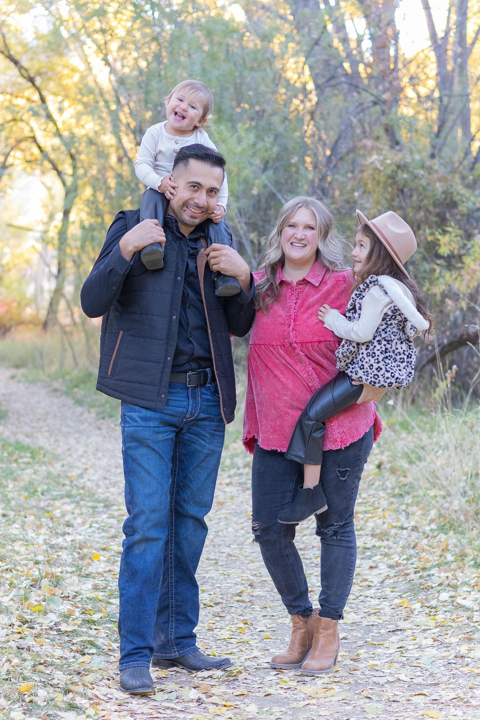 A family stands in the woods in the fall, with mom tickling the baby, who is on the dad's shoulders. The oldest daughter is smiling at  the baby's joy. Outdoor family sessions by the Lens Lady are a fun way to make special memories as a family.