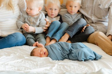 A newborn baby lies on the bed in front of his three older brothers and mom and dad. The Lens Lady can capture these precious days in the comfort of your own home.