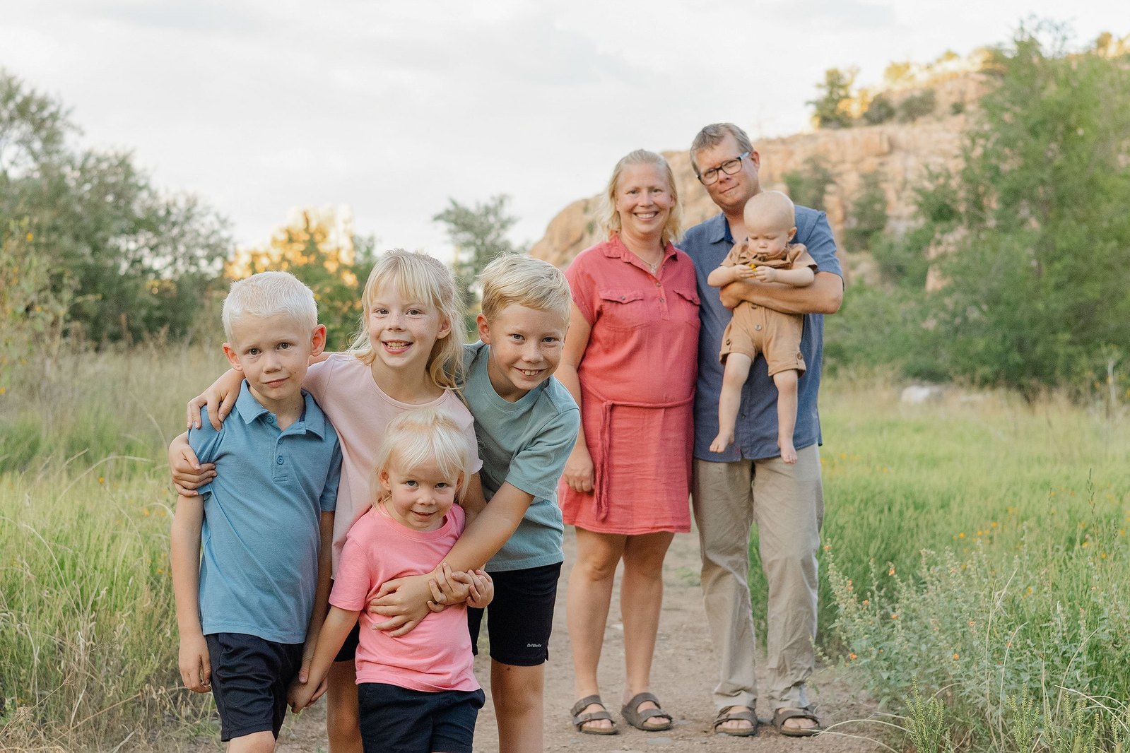 Four children hugging and grinning at the camera while mom and dad stand arm in arm in the distance with the youngest in their arms. This fun, summer family portrait was taken by Keilah Johnson, the Lens Lady.