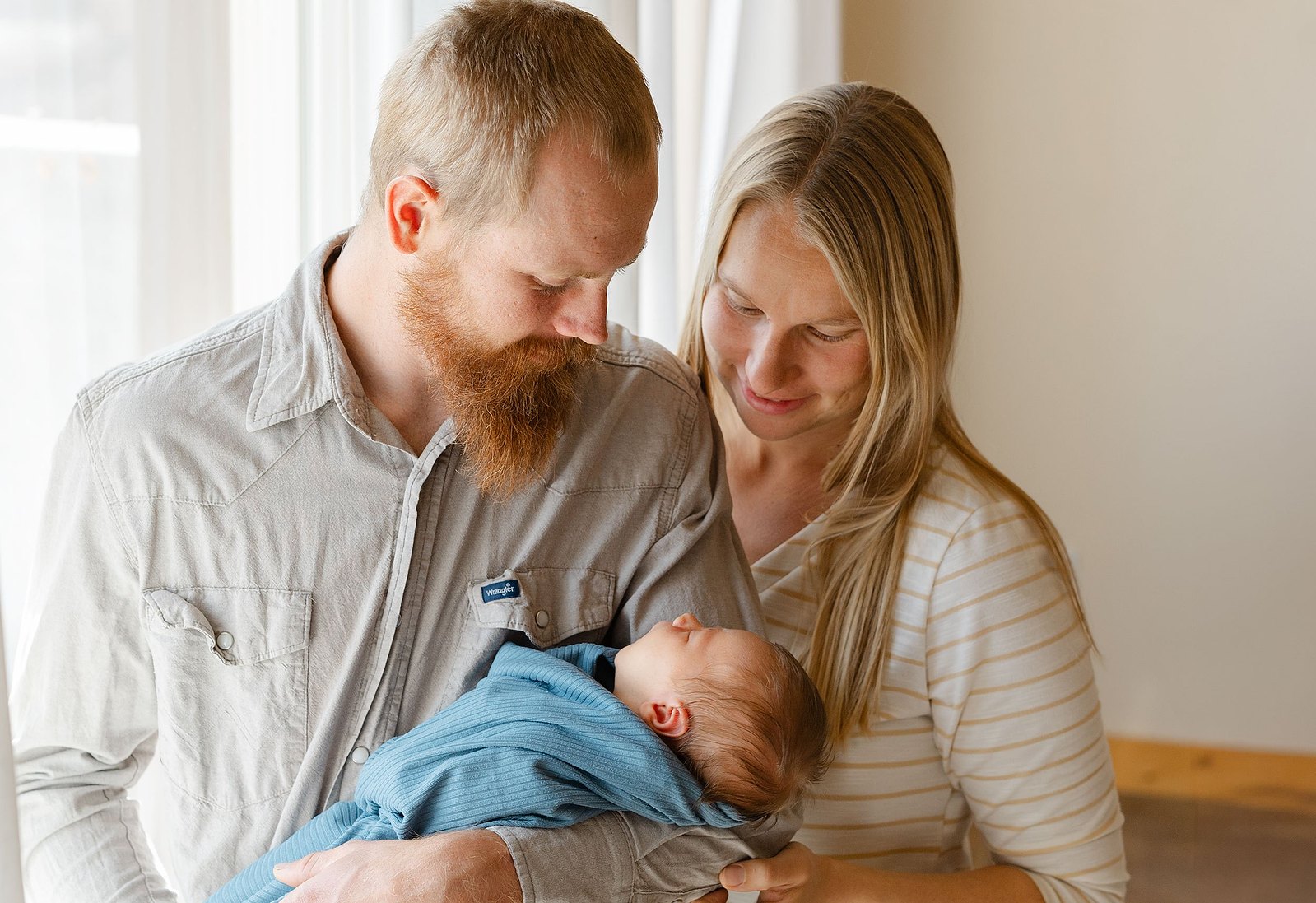 A new mom and dad hold their baby boy and look at him with love in a bright and airy room with natural light, photographed by Keilah Johnson, the Lens Lady.
