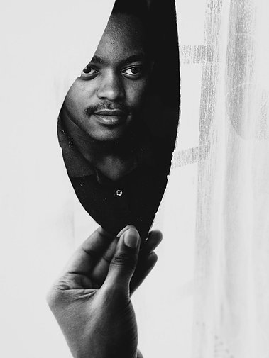 La main d'un homme noir tient un morceau de miroir dans lequel se reflète son visage. Face à lui une fenêtre couverte d'un voile blanc. La photo est en noir-et-blanc.