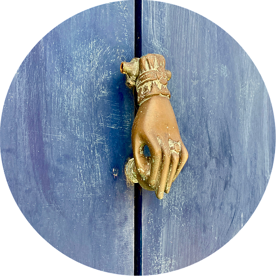 Sur une porte en bois lazurée en bleu se trouve un heurtoir doré en forme de main fine invitant la personne à frapper à la porte.