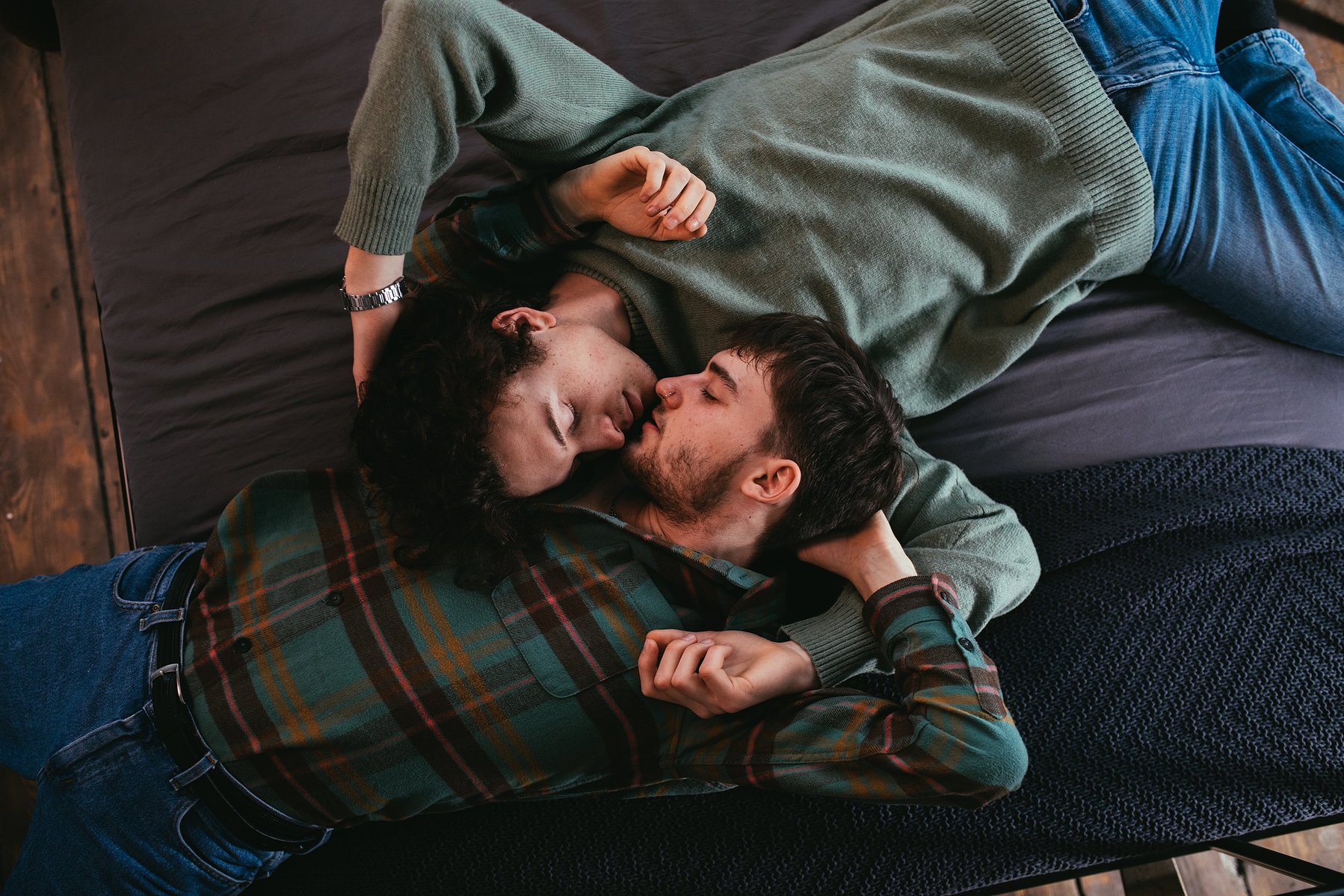 Un couple est allongé en quinconce sur un sofa mou et s'embrasse avec douceur. L'homme et la femme sont dans la trentaine, châtains foncés tous les deux, blancs, et portent des vêtements similaires aux teints verts et bleus. 